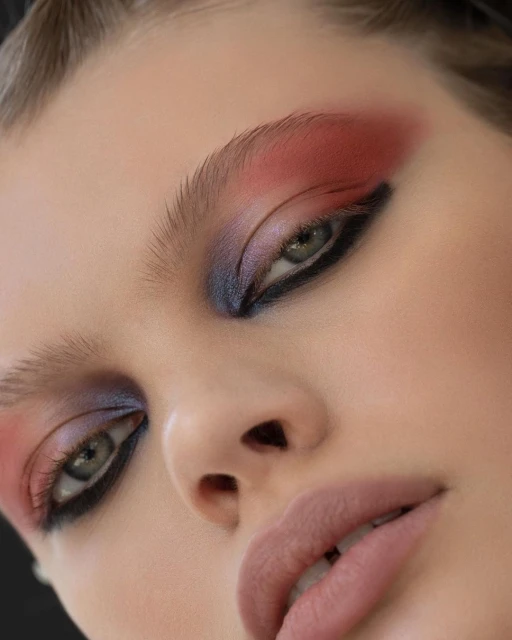 Close-up portrait of a woman's face featuring vibrant and dramatic makeup with red, purple, and black eyeshadow and neutral lipstick.