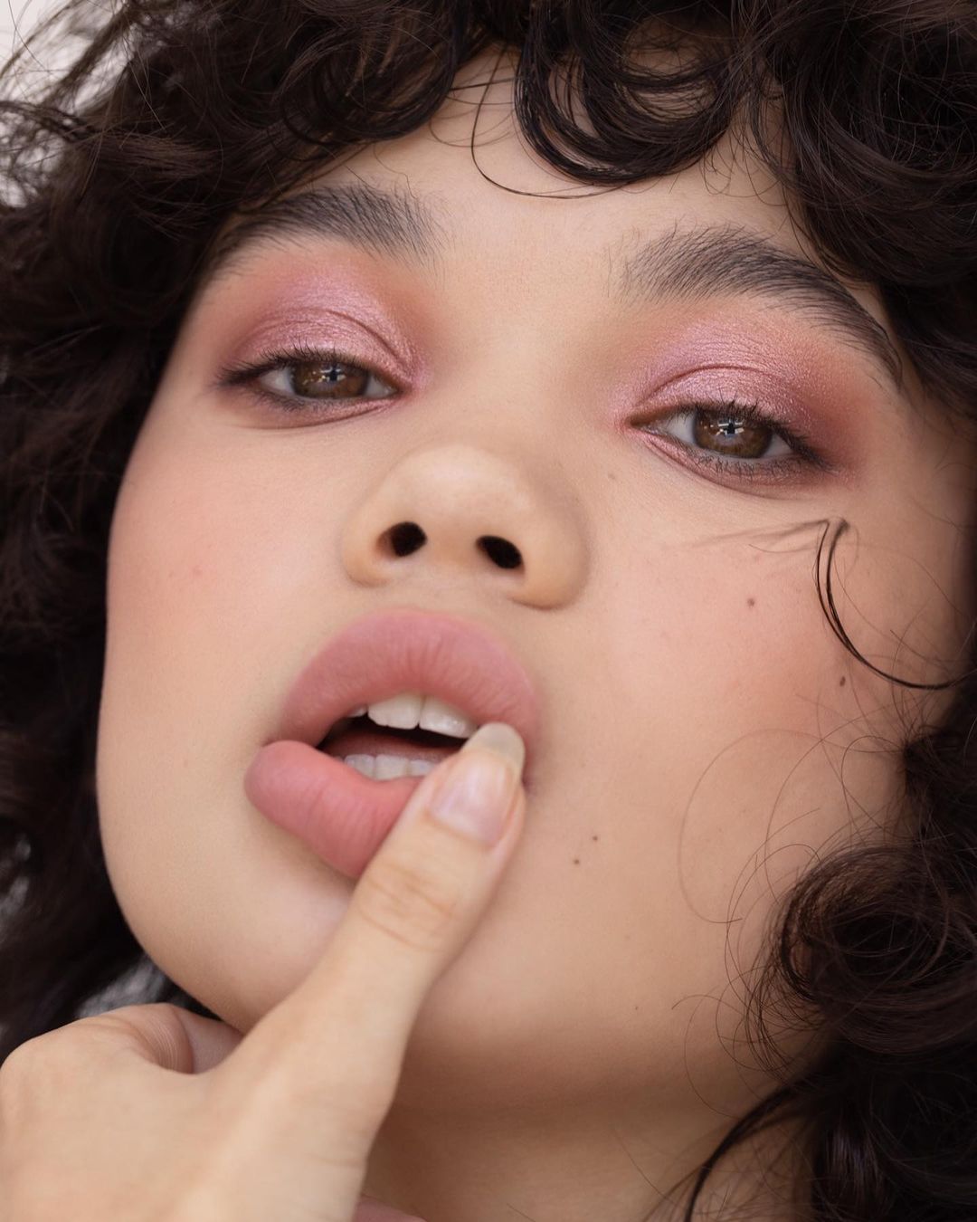 Close-up portrait of an adult female with pink eyeshadow, lips slightly open, and a finger near her mouth in a beauty style.