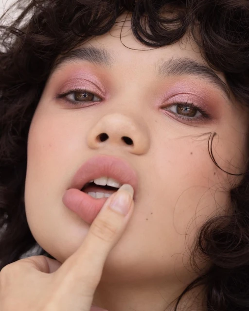Close-up portrait of an adult female with pink eyeshadow, lips slightly open, and a finger near her mouth in a beauty style.