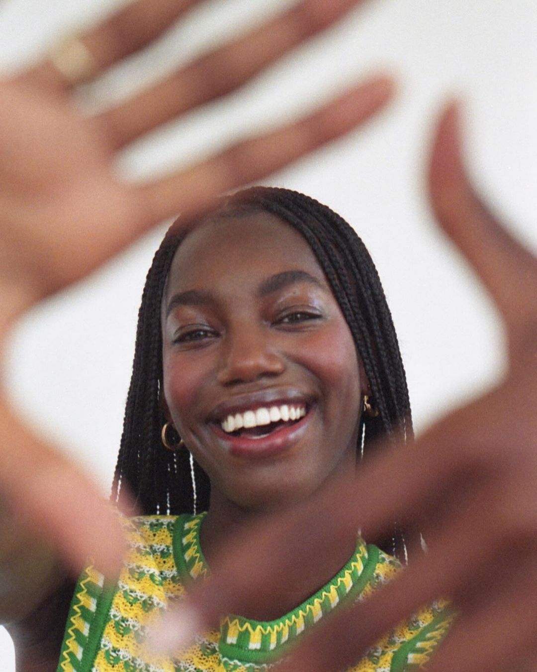 Smiling African-American woman with braids frames her face with her hands, wearing a yellow and green sleeveless top against a white background.