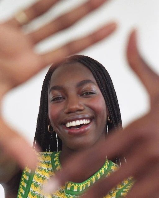 Smiling African-American woman with braids frames her face with her hands, wearing a yellow and green sleeveless top against a white background.