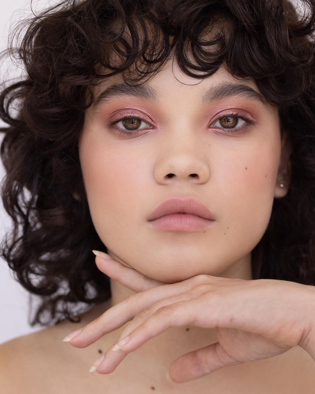 Close-up portrait of a young adult female with curly hair, light skin, and pink eye shadow posing in front of a white background.