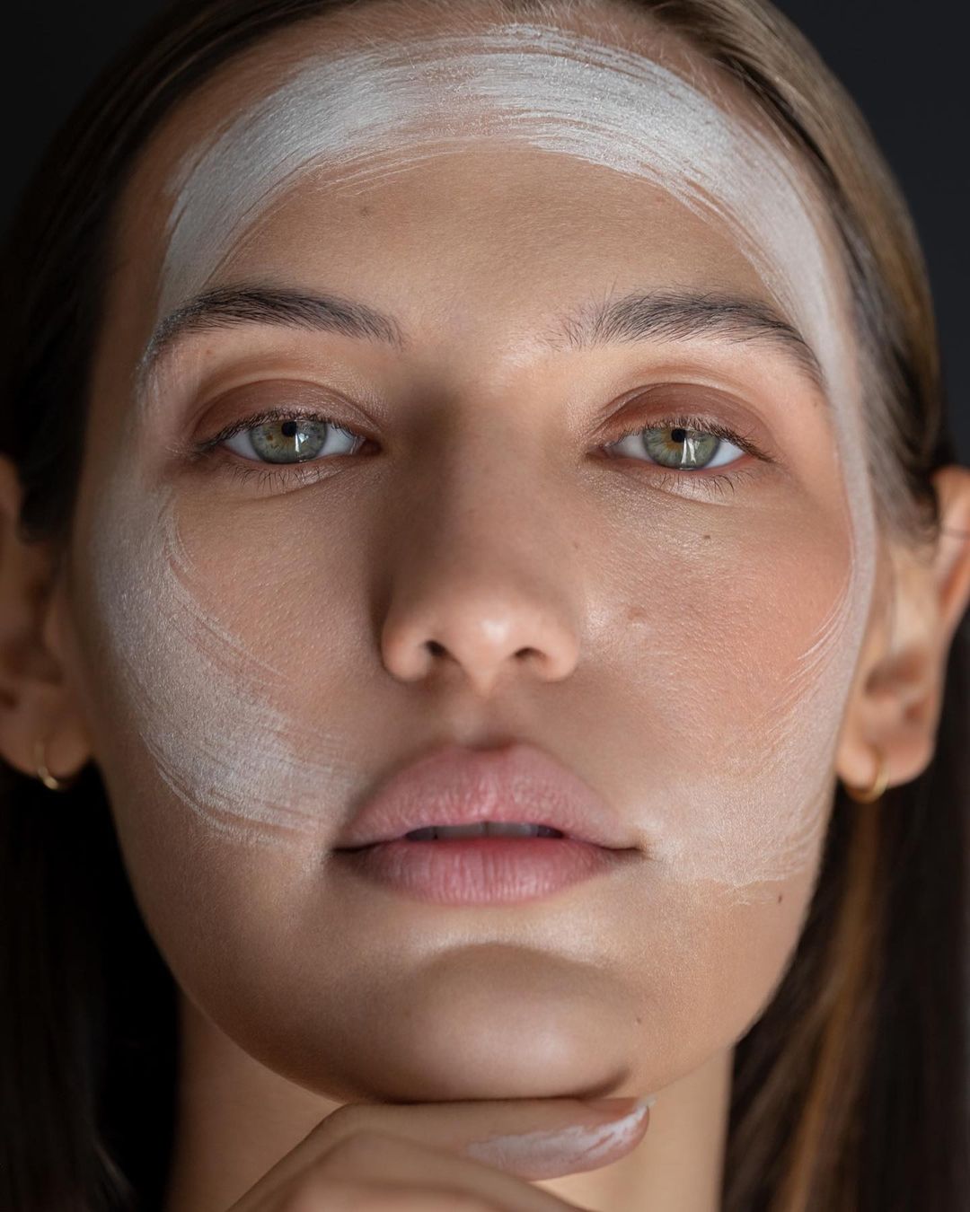 Close-up portrait of a woman with facial mask, showing skincare or beauty treatment, against a dark neutral background in soft lighting.