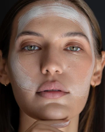 Close-up portrait of a woman with facial mask, showing skincare or beauty treatment, against a dark neutral background in soft lighting.