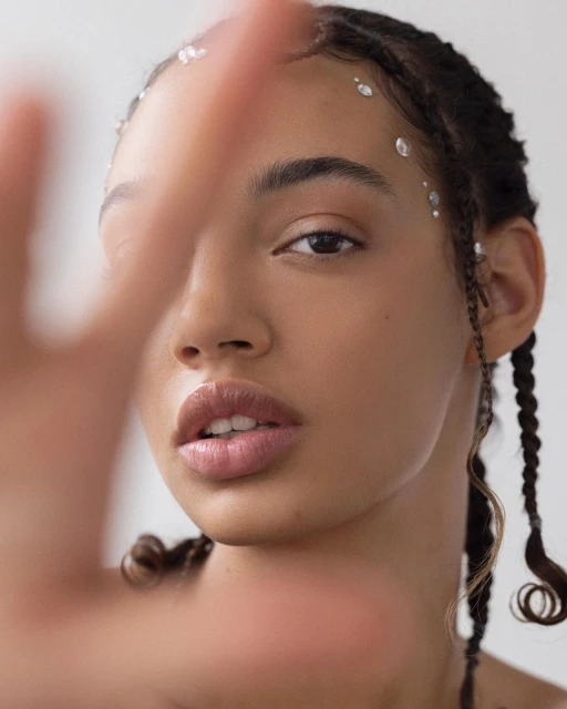 Close-up portrait of a young woman with dark hair styled in braids, accented with small jewels, posing with hand partially obscuring face.