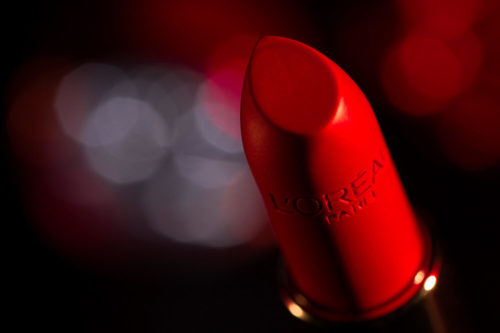 Macro shot of vibrant red lipstick tube with visible 'L'Oreal Paris' brand, set against soft, blurred lights creating a beauty product advertisement.