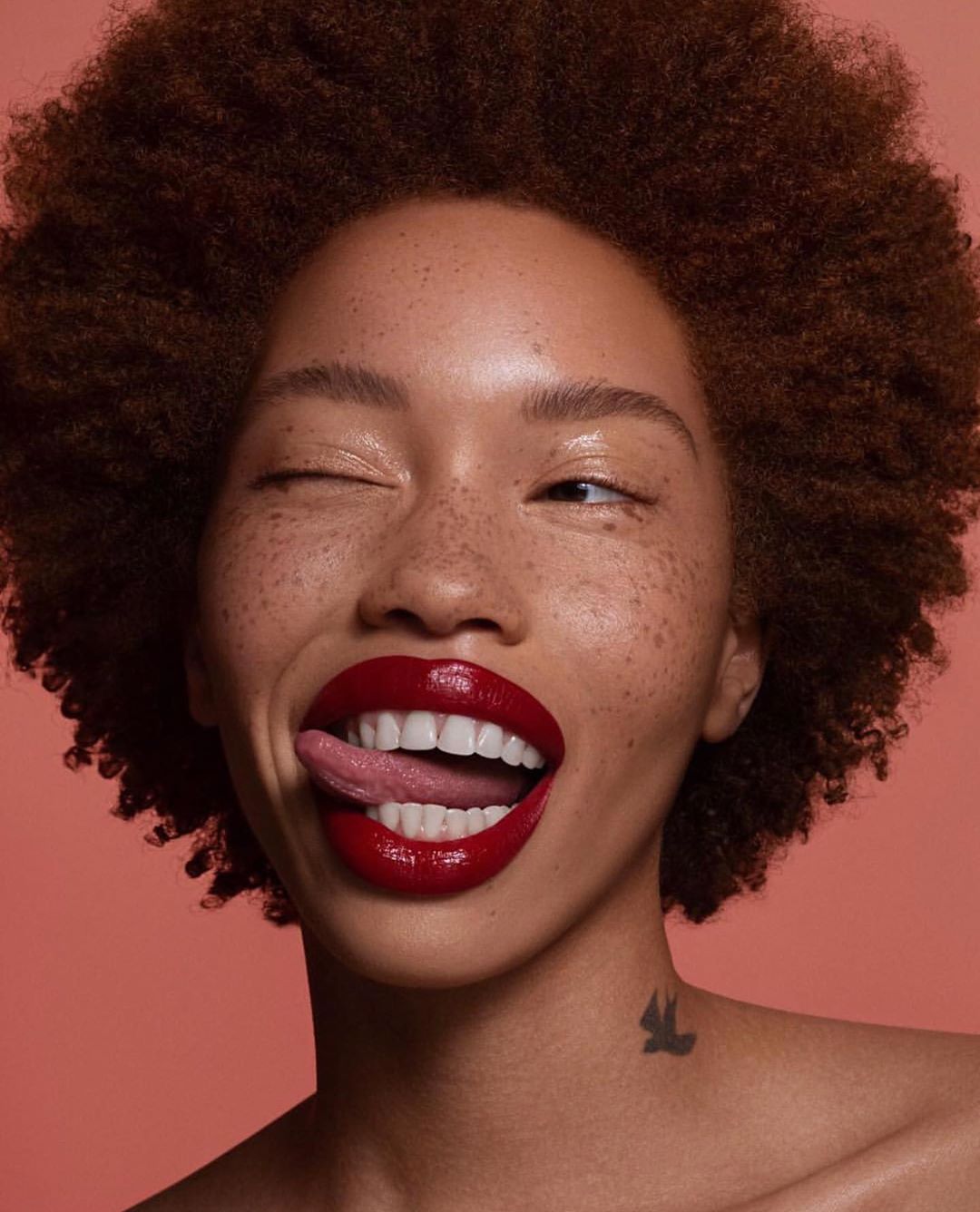 Portrait of a young adult woman with an afro and freckles sticking out her tongue, wearing red lipstick, against a salmon pink background.