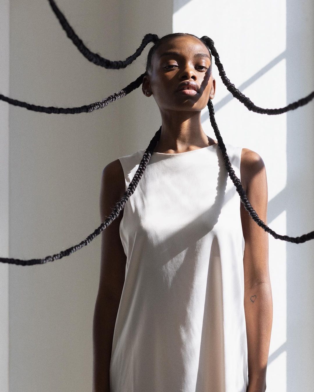 Portrait of a Black woman in a white sleeveless top with long black braided extensions stringing outwards against a white wall.