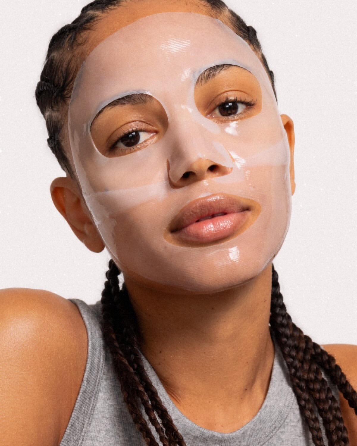 Close-up of a woman with braided hair wearing a sheet mask, showcasing beauty and skincare. Shot against a neutral backdrop.