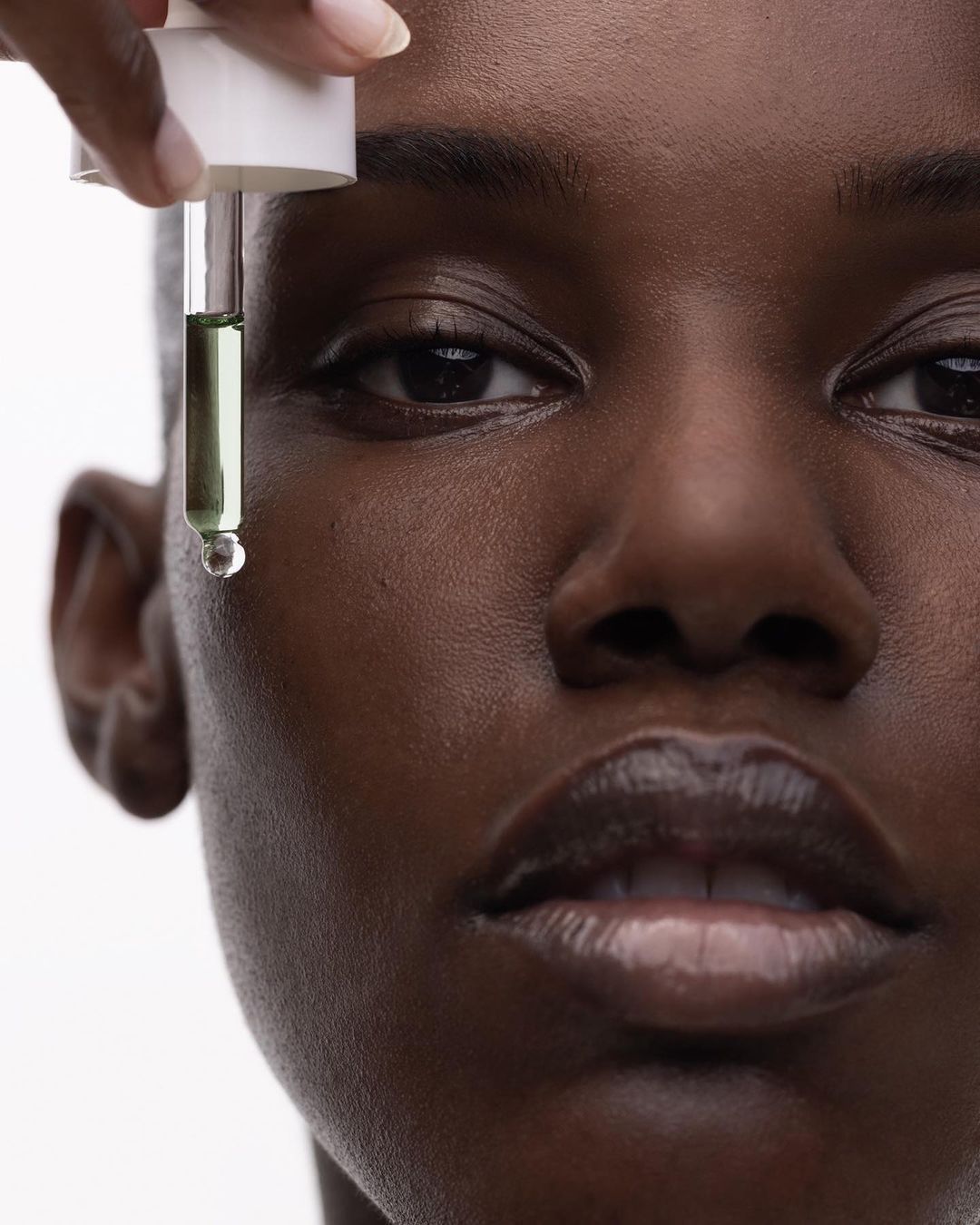 Close-up shot of a dark-skinned adult woman's face, with a dropper applying facial oil. Focus on beauty and skincare product.