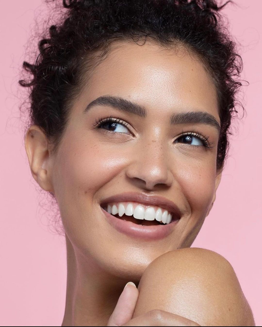 Close-up studio portrait of an adult woman smiling, looking to the side, and showing white teeth against a light pink background.