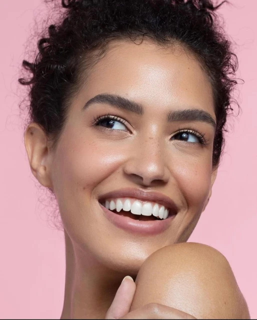 Close-up studio portrait of an adult woman smiling, looking to the side, and showing white teeth against a light pink background.