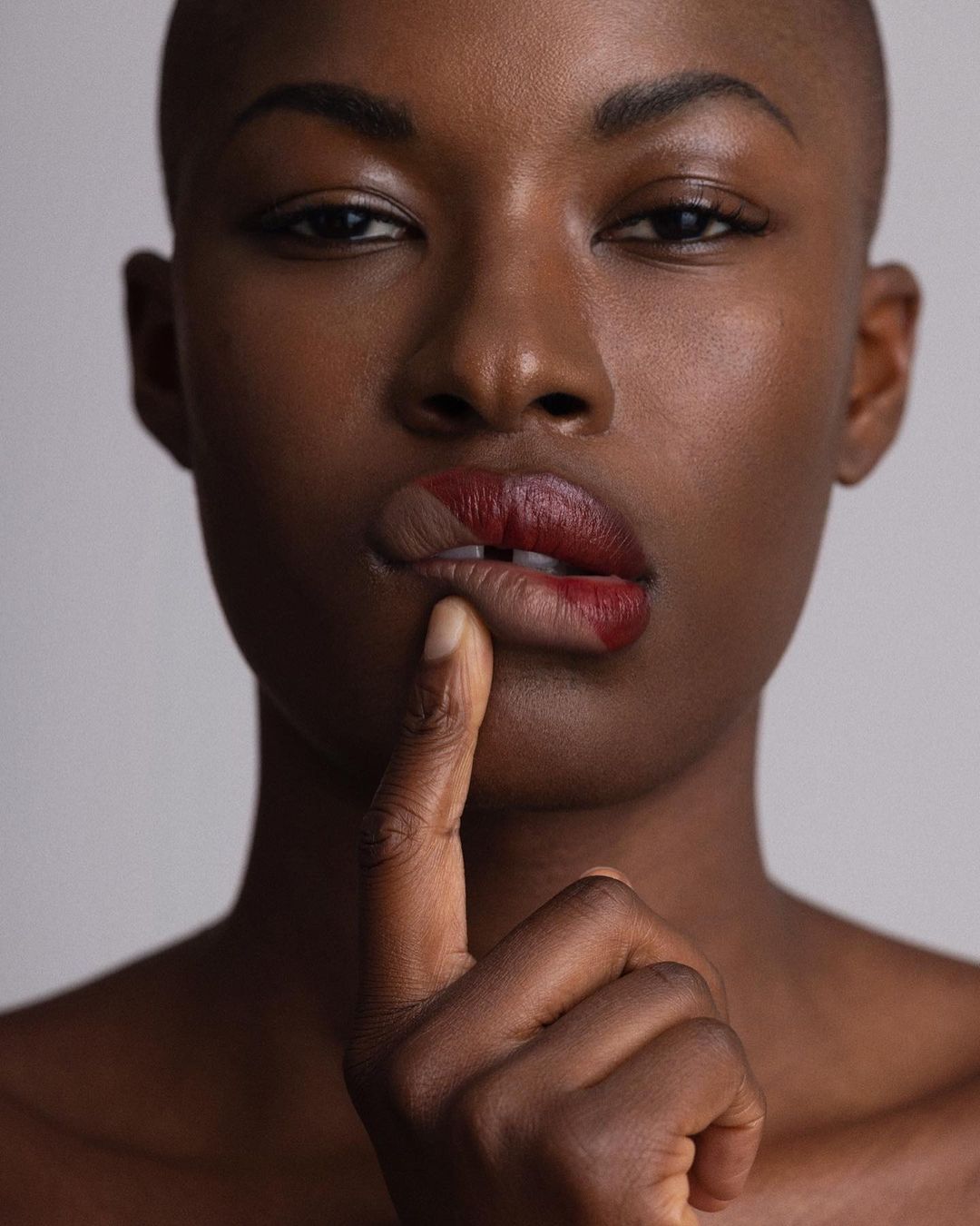 Close-up of a Black female adult with bare shoulders and a shaved head, with a finger held to her lip in a studio setting.