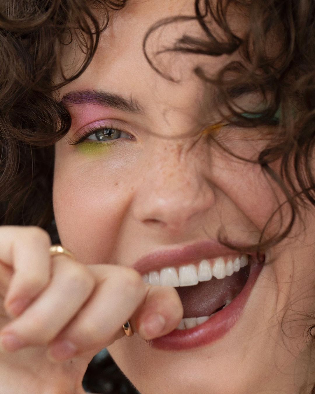 Close-up portrait of a smiling young woman with curly brown hair and vibrant makeup, posing with her hand near her mouth indoors.