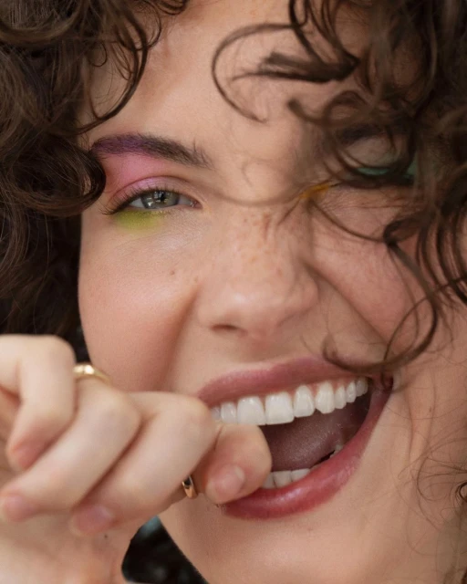 Close-up portrait of a smiling young woman with curly brown hair and vibrant makeup, posing with her hand near her mouth indoors.