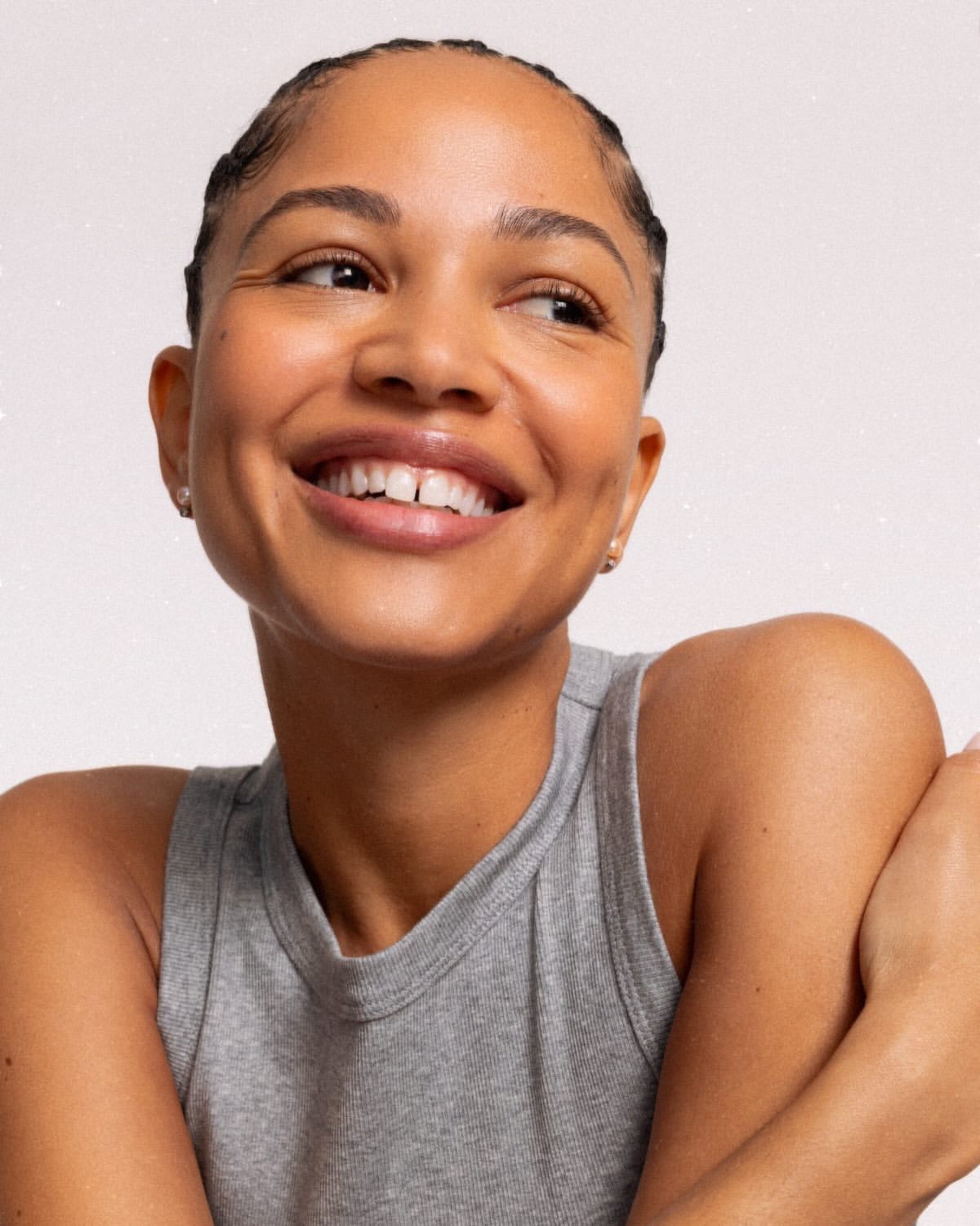 Close-up of a smiling woman with braided hair wearing a gray tank top, set against a white background, showcasing a beauty portrait.