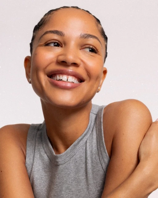 Close-up of a smiling woman with braided hair wearing a gray tank top, set against a white background, showcasing a beauty portrait.