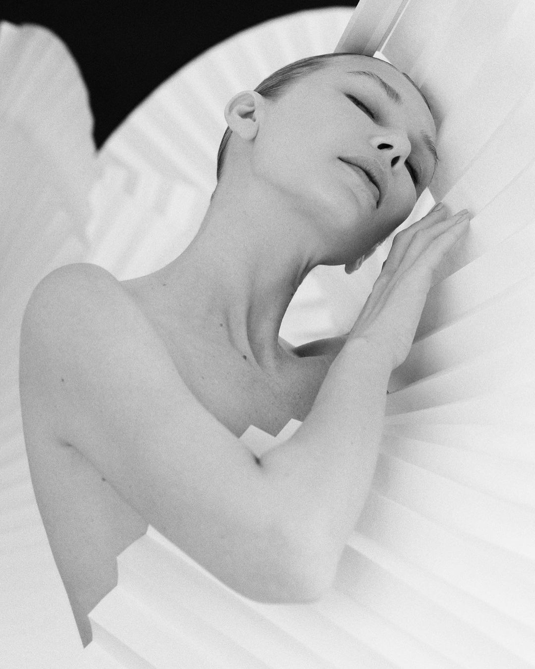 Black and white portrait of a woman with closed eyes resting her arm against a pleated fan-like background element in an indoor space.