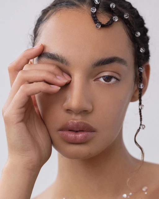 Close-up studio shot of a woman with braided hair adorned with clear beads, fair skin, and light makeup against a neutral background.