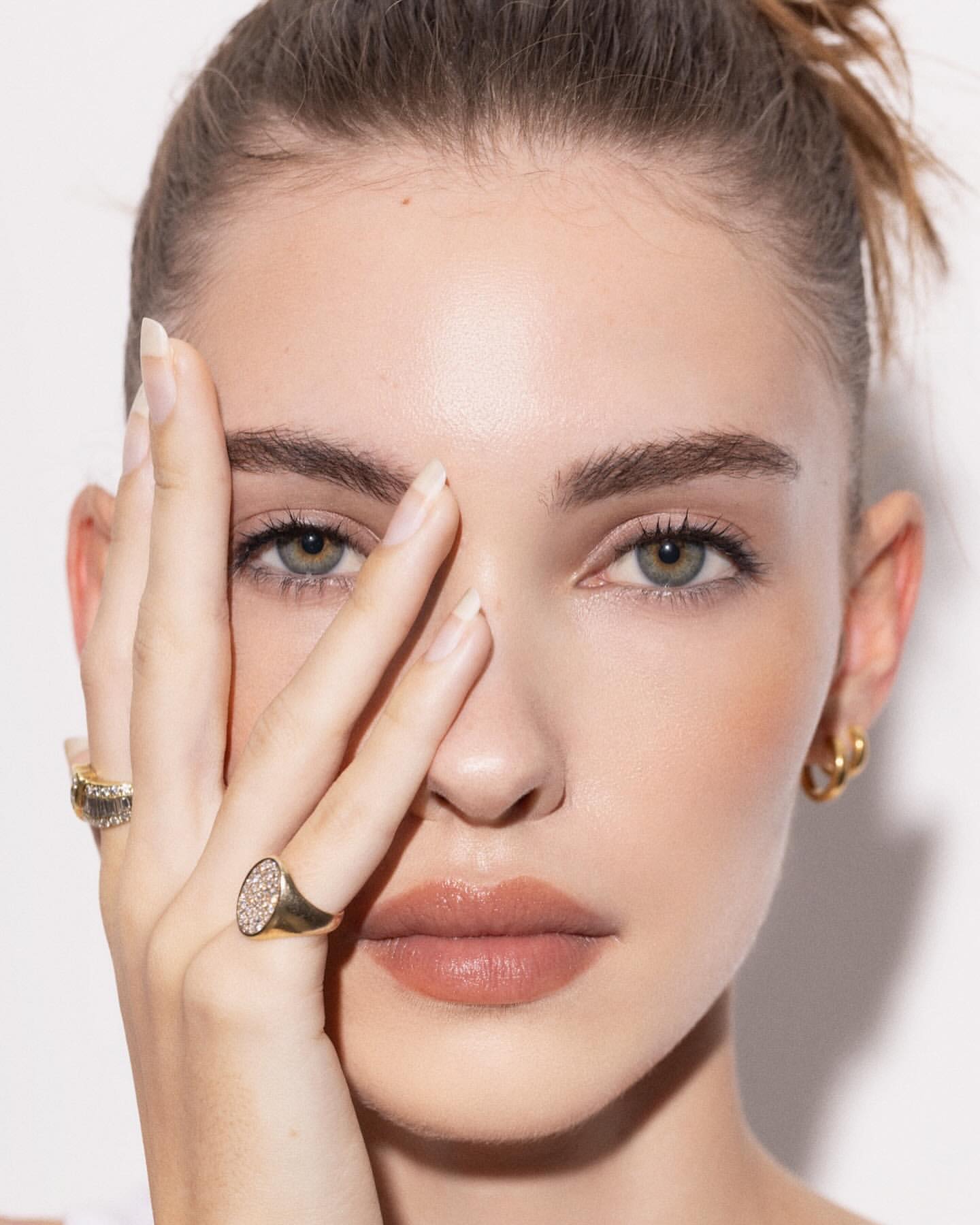Close-up portrait of an adult woman with fair skin, her hand partially covering her face, accessorized with rings, set against white background.