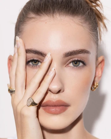 Close-up portrait of an adult woman with fair skin, her hand partially covering her face, accessorized with rings, set against white background.