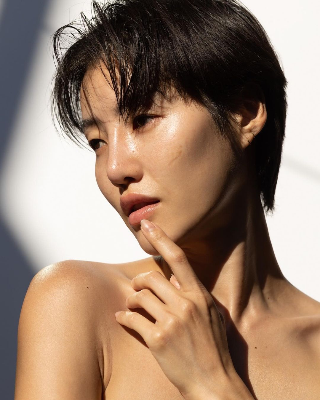 Close-up studio shot of a woman with short dark hair. She has bare shoulders, a light complexion, and she is touching her face with her fingers.