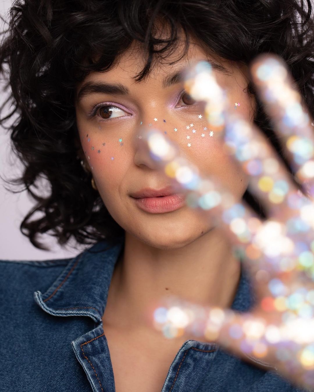 Close-up portrait of a woman with dark curly hair, star glitter on her face, and a hand with bokeh sparkles in the foreground.