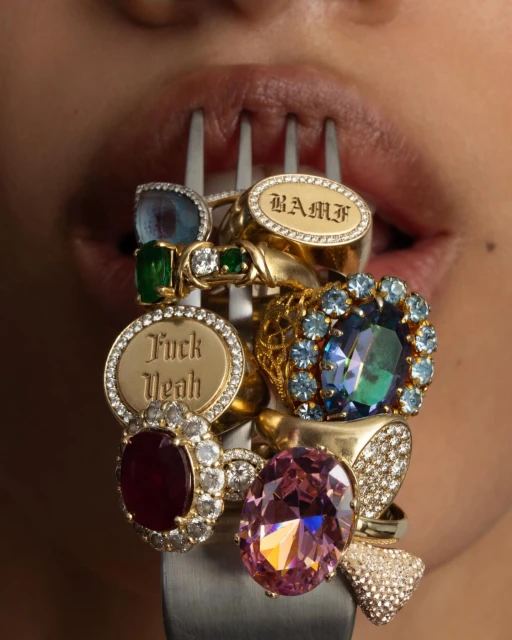 Close-up shot of a person holding a fork laden with numerous gold rings set with various gemstones and inscribed text.