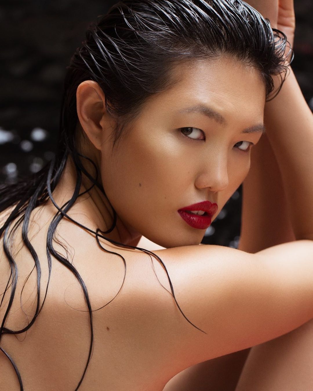 Close-up portrait of a woman with wet, dark hair, red lipstick, and bare shoulders against a dark, blurred background.