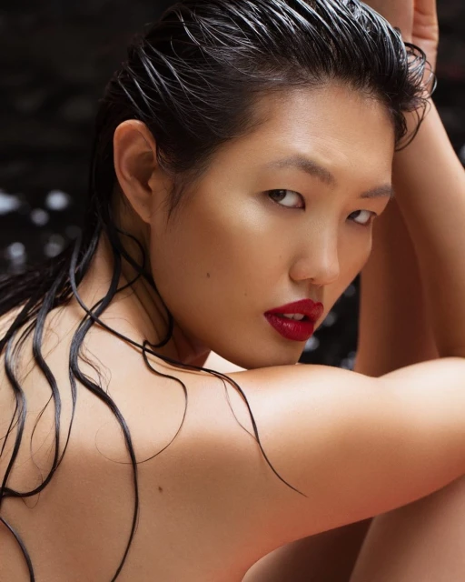Close-up portrait of a woman with wet, dark hair, red lipstick, and bare shoulders against a dark, blurred background.