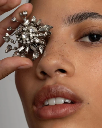 Close-up portrait of a woman holding a large crystal-encrusted brooch over her right eye by Angela Marklew