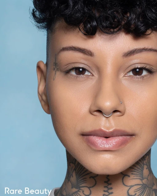 Close-up portrait of an adult woman with tattoos and short curly hair against a light blue background, showcasing beauty and personal style.