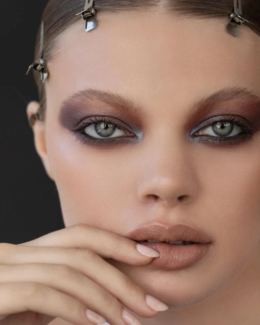 Close-up portrait of a young woman with dramatic eye makeup, manicured nails, and hair styled with metal clips against a dark background.