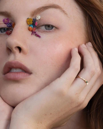 Close-up portrait of a woman with colorful dried flowers glued around her eyes and a thin gold ring on her hand, photographed by Angela Marklew.