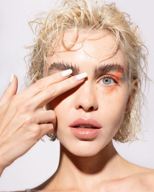 Close-up of a woman with blonde hair and bright makeup, covering one eye with her hand, against a plain white background.
