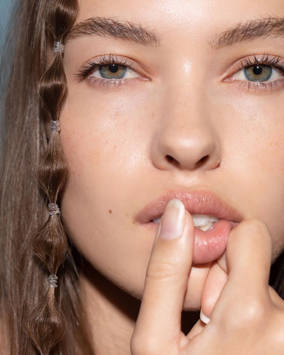Close-up of a woman's face with brown hair styled in bubble braids, light skin, and blue-green eyes, soft lighting, beauty concept.