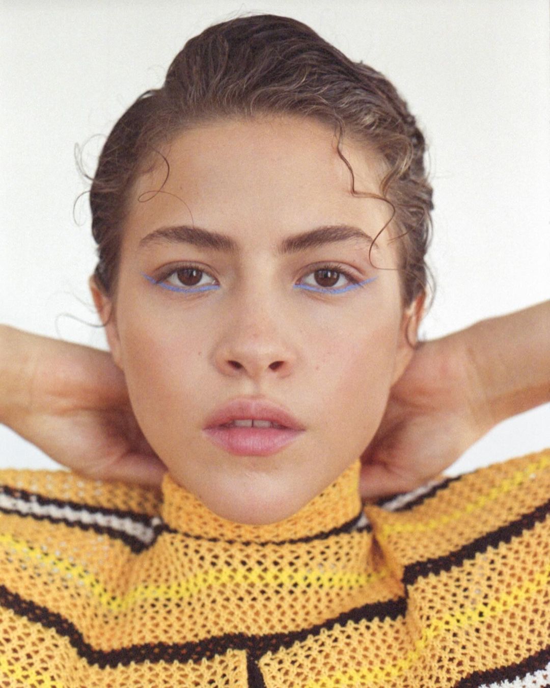 Close-up of a woman with slicked-back brunette hair and blue eyeliner, wearing a yellow and black striped knit top against a white backdrop.