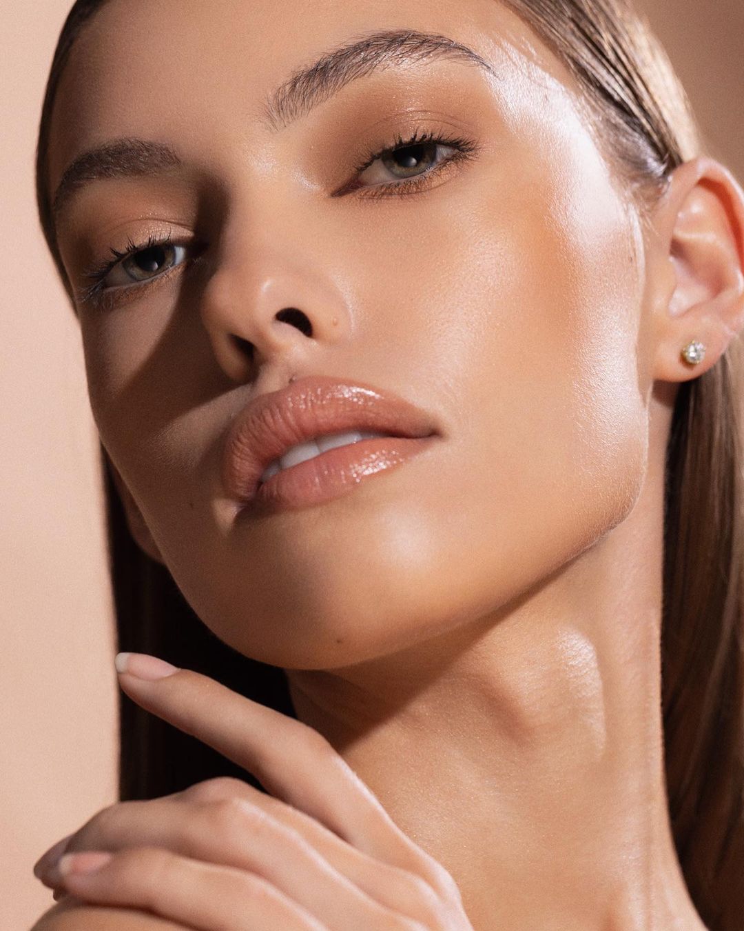 Close-up portrait of a young adult woman with slicked-back hair and glossy makeup against a neutral backdrop in a beauty-style photograph.