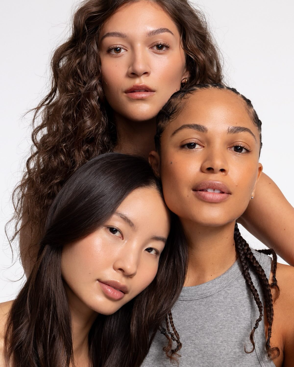 Close-up studio portrait of three diverse young adult women with natural makeup and hairstyles against a bright white backdrop.