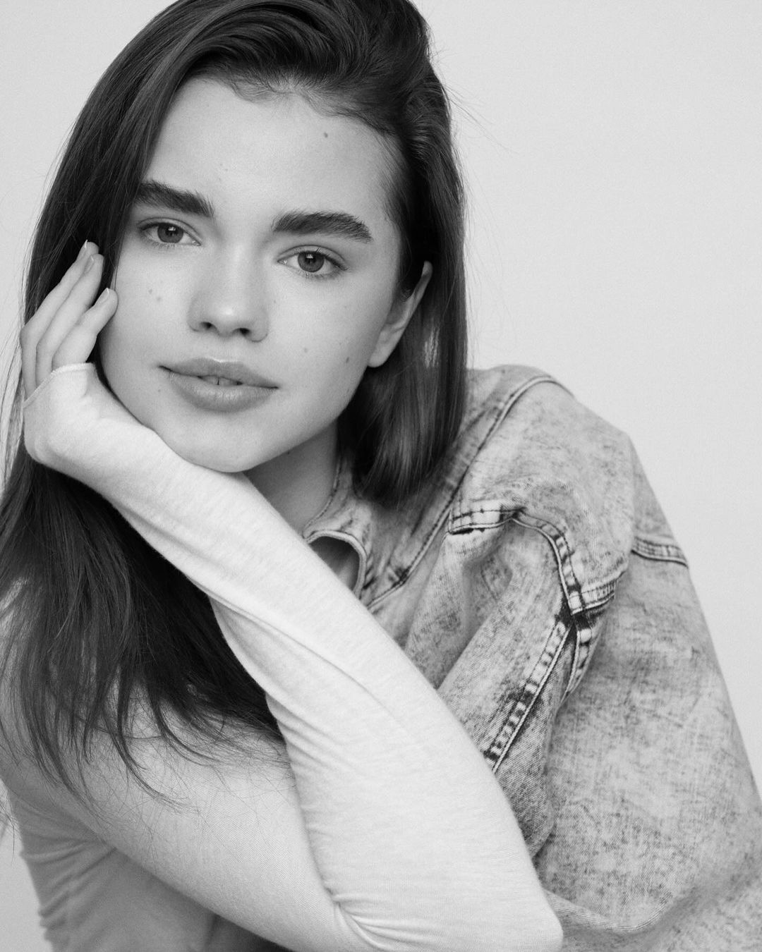 Black and white portrait of a young adult woman with hand near her face, wearing a denim jacket against a plain background.