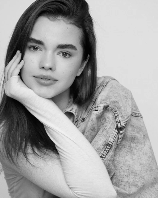 Black and white portrait of a young adult woman with hand near her face, wearing a denim jacket against a plain background.