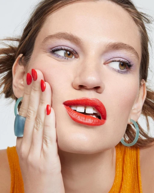 Close-up of a woman with red lipstick, light purple eye shadow and an orange top posing in front of a white background.