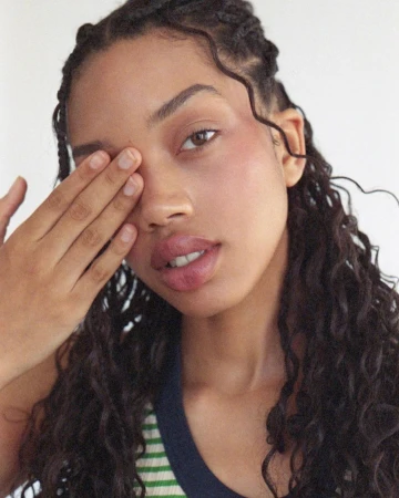 Close-up portrait of a young woman with brown curly hair covering one eye and looking directly at the camera against a white background.