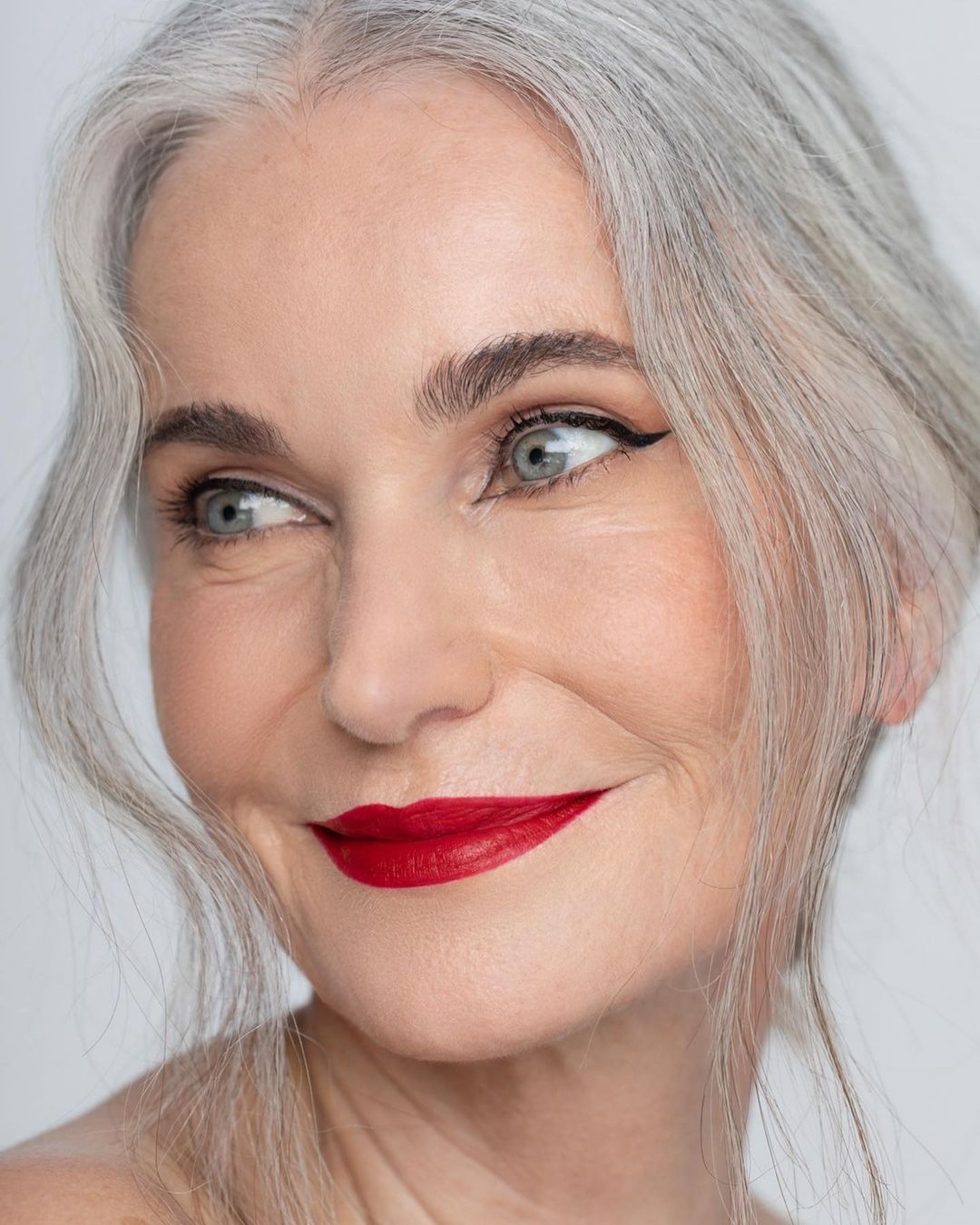 Close-up portrait of a mature woman with silver hair, bright red lipstick, and eye liner, looking off to the side against a white background.