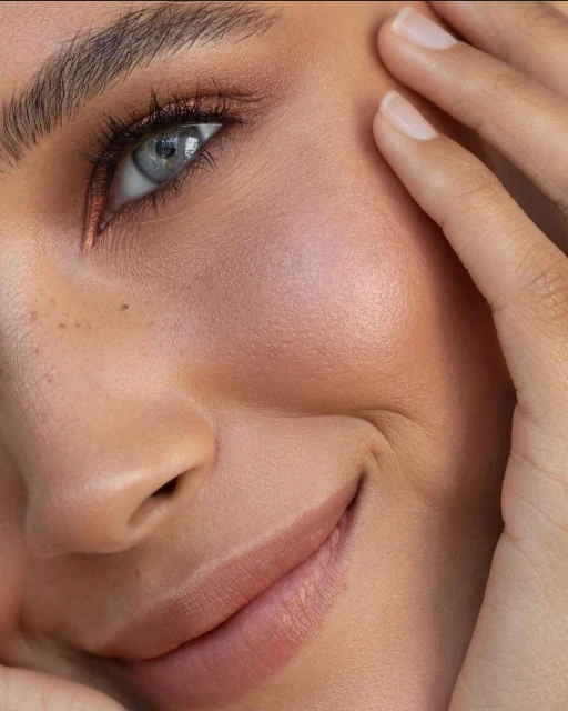Close-up portrait of an adult female's face showcasing makeup, with focus on her eye, cheek, and lips. Hands are gently touching her face.