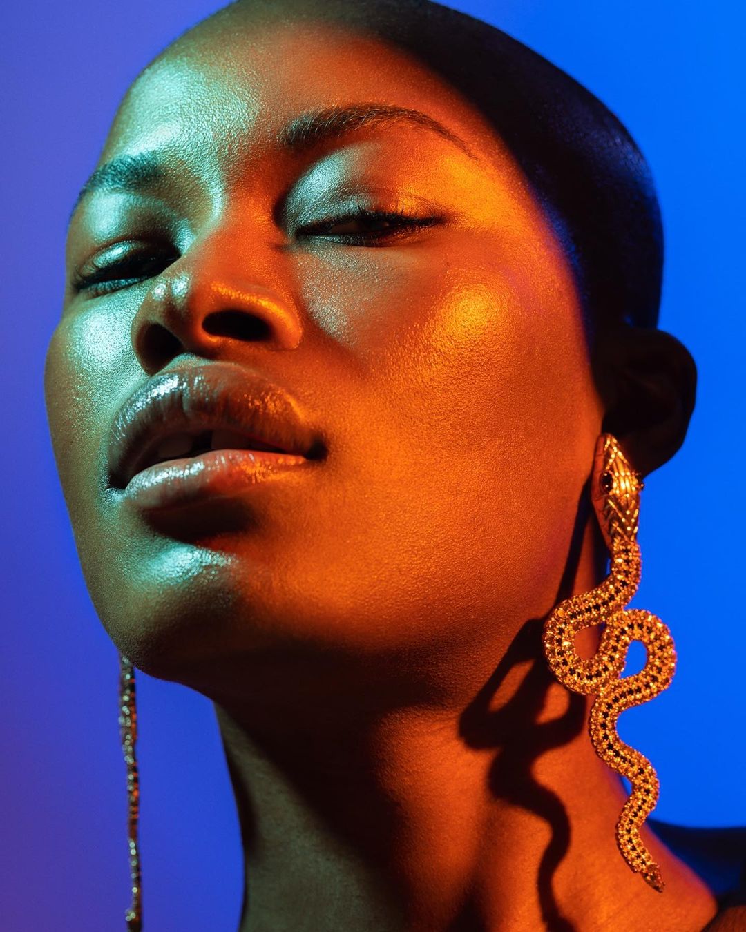Close-up portrait of a Black woman with closed eyes, orange-gold lighting, and snake earrings against a vibrant blue background.