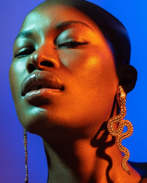 Close-up portrait of a Black woman with closed eyes, orange-gold lighting, and snake earrings against a vibrant blue background.