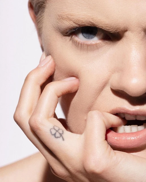Close-up of a woman with fair skin, blue eyes, and a hand near her face. She has a tattoo of a clover on her finger.