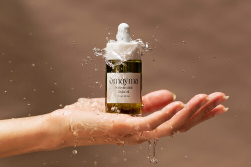 Product shot of Omoyma Bedouin Elixir facial oil on a woman's palm with water splashing against a neutral tan background.