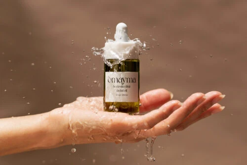 Product shot of Omoyma Bedouin Elixir facial oil on a woman's palm with water splashing against a neutral tan background.
